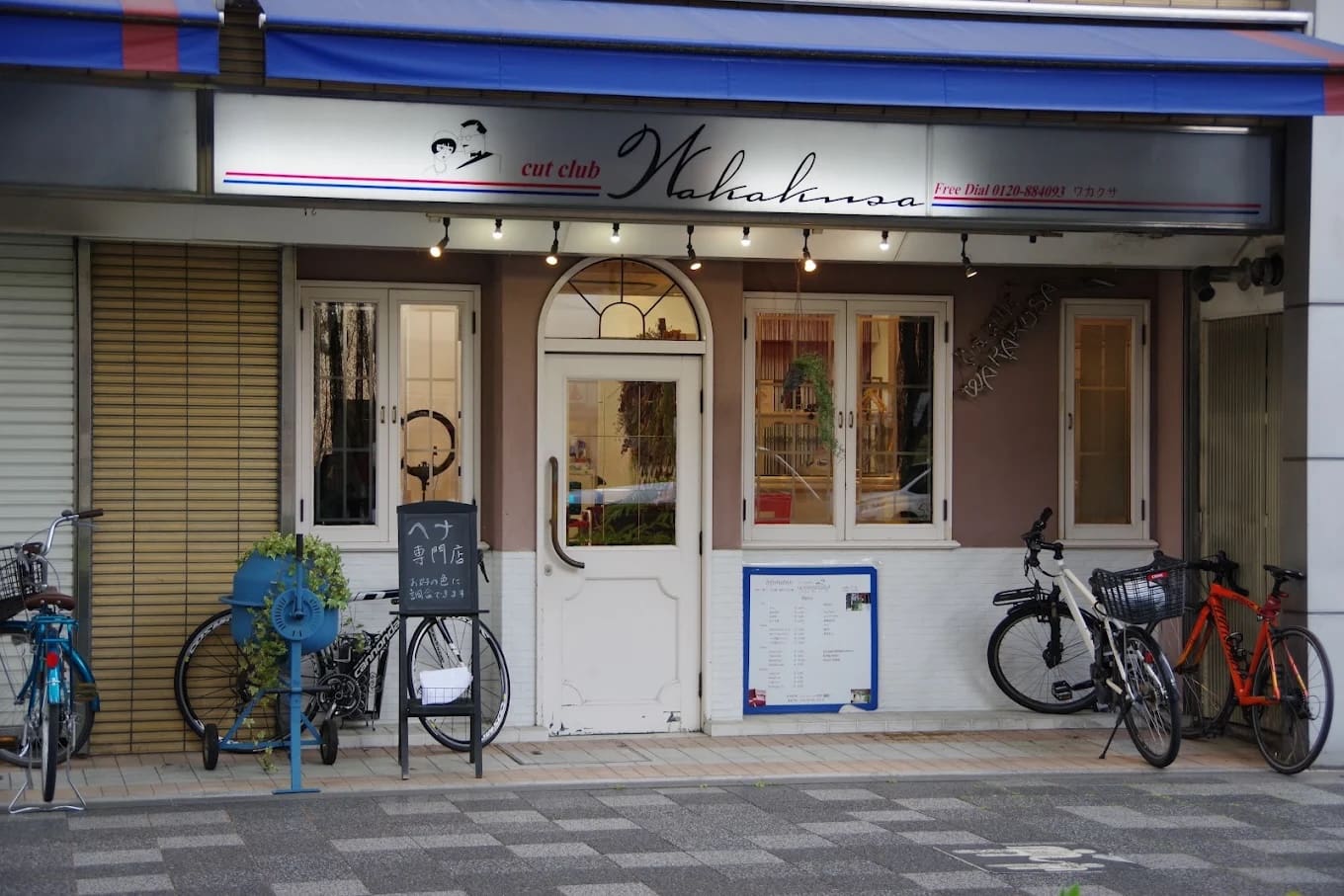 A loja tem uma placa que diz "cut club Wakana" em cima de uma porta dupla branca e oferece serviços de atalho e henna. Imprensado entre as janelas e o estacionamento de bicicletas, há bicicletários azuis, plantas de casa, menus e placas perto da entrada.