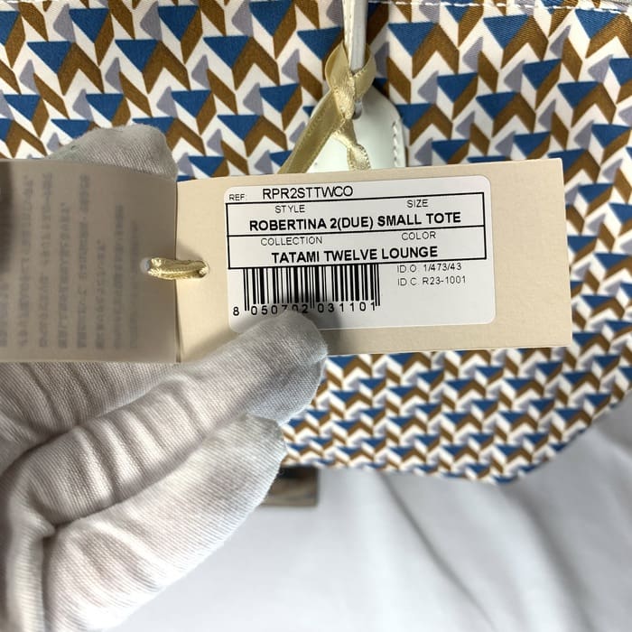 A mão enluvada segura a etiqueta do produto "ROBERTINA 2 (DUE) SMALL TOTE" na parte superior do tecido com padrão de tatame com um padrão geométrico em azul, marrom e branco, que é o estilo de assinatura da coleção de pequenas bolsas ROBERTA PIERI.