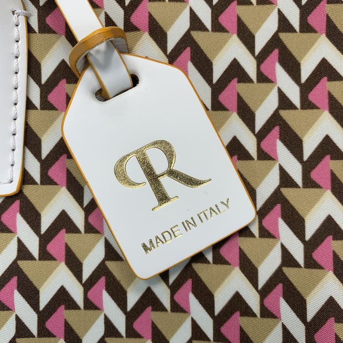 Um close-up de uma etiqueta de bagagem branca Roberta Pieri com as letras "R" em ouro e as palavras "MADE IN ITALY" presas a uma mini mochila com padrões geométricos marrons, bege e rosa.