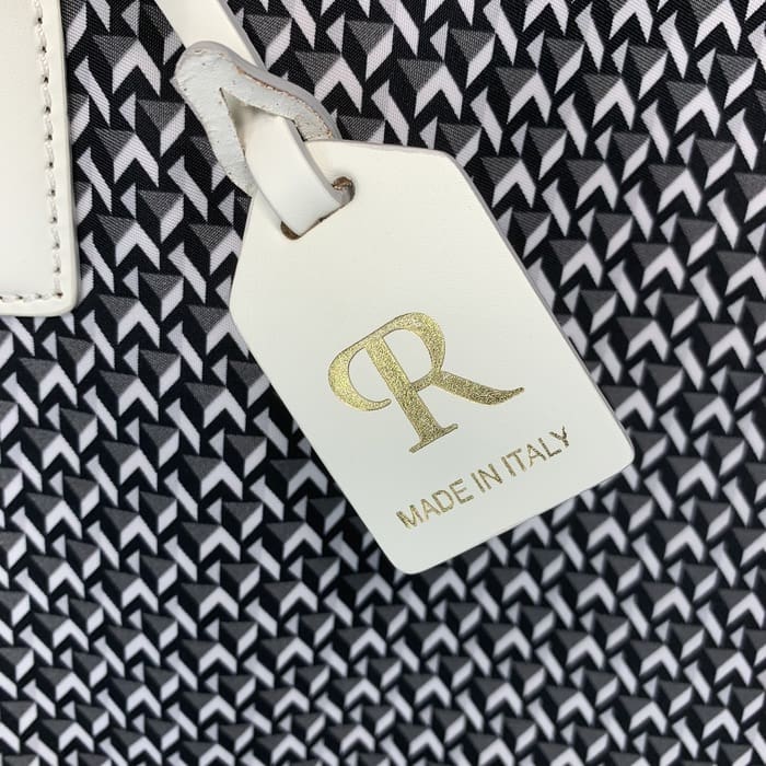 Uma etiqueta ROBERTA PIERI de couro branco com as letras "R" em ouro e as palavras "MADE IN ITALY" está pendurada no fundo de um tecido geométrico preto e branco.