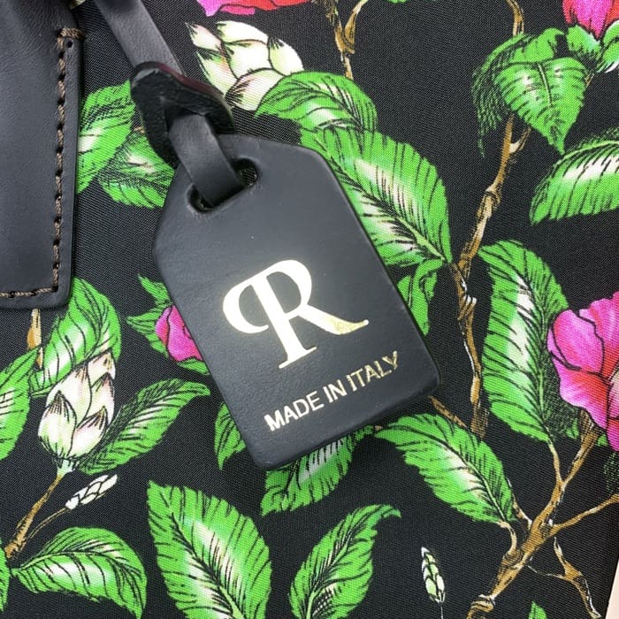 A etiqueta de bagagem preta ROBERTA PIERI com o "R" dourado e texto italiano é suspensa em um tecido floral com folhas verdes e flores rosa, complementando a mini mochila com elegância italiana atemporal.