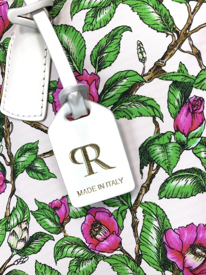 Grande plano da mini mochila ROBERTA PIERI. pedúnculos com folhas verdes e flores rosa; A etiqueta branca anexada à bolsa de 2 vias apresenta um logotipo "R" dourado e as palavras "MADE IN ITALY".